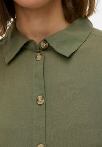 Camicia verde oliva con colletto in tessuto texturizzato, caratterizzata da sei bottoni beige disposti sul davanti, che mostrano un design semplice e classico.