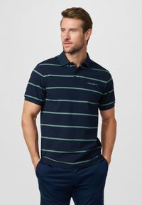 Mann mit kurzen Haaren, trägt ein marineblau-grün gestreiftes Poloshirt mit "Hackett"-Logo, Hände in den Taschen, schaut seitlich vor einfachem Hintergrund.