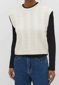 Pull sans manches tricoté de couleur crème avec ourlet côtelé et motif torsadé, porté sur un haut à manches longues noir et associé à un jean bleu.