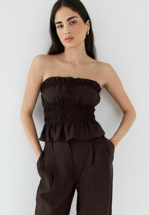 Junge Frau mit langen dunklen Haaren, die ein trägerloses schwarzes Top mit Raffung und passende hoch taillierte Hose trägt, steht mit Händen in den Taschen.