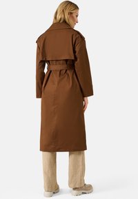 Cappotto trench marrone con vita alta e cinturo, maniche lunghe e dettaglio sul retro. Indossato sopra pantaloni larghi beige chiaro e scarpe chunky beige.