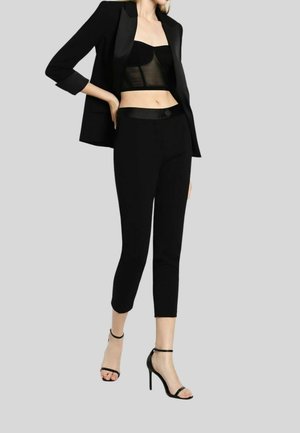 Kvinde iført sort blazer, gennemsigtigt cropped bustier, cropped sorte bukser og sorte højhælede sko med ankelrem mod en grå baggrund.