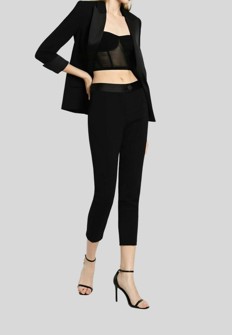 Femme portant un blazer noir, un bustier court transparent, un pantalon noir court et des talons hauts noirs à bride cheville sur fond gris.
