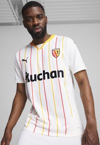 Camiseta deportiva blanca con rayas verticales rojas y amarillas, con cuello en V, logo de Puma, texto negro "Auchan" y un escudo de club.