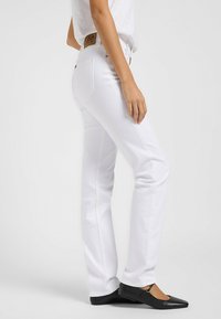 Jeans rectos de mezclilla blanca con bolsillos traseros visibles, detalles de costura y un parche de logo de cuero en la cinturilla. Zapatos planos negros incluidos.