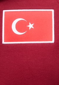 Κόκκινο ύφασμα με ραμμένη ορθογώνια επιφάνεια που απεικονίζει τη τουρκική σημαία: λευκό ημισέληνο και αστέρι σε κόκκινο φόντο με λευκή περίμετρο.