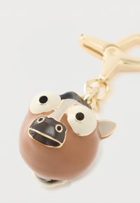 Barna és fekete, rajzfilmes stílusú kutyás kulcstartó nagy, kerek szemekkel és aranyszínű kapoccsal, fehér háttéren.