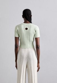 Kobieta z warkoczem, ubrana w jasnozielony, prążkowany top z krótkim rękawem, z okrągłym wycięciem z tyłu i kremowymi koronkowymi spodniami.