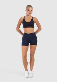 Zwarte sportbh met plooidetails en verstelbare bandjes, gecombineerd met strakke marineblauwe shorts. Model draagt witte sportschoenen.
