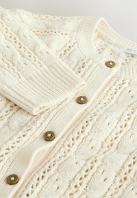 Cream cable-knit cardigan featuring ribbed cuffs and six brown buttons, displaying intricate knitted patterns and a soft texture.