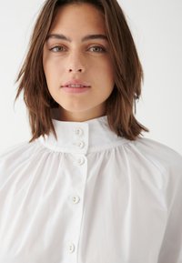 Weiße, kurzärmelige Bluse mit hohem Kragen, gerafften Details und glatter Stofftextur. Verfügt über klare, runde Knöpfe an der Vorderseite.