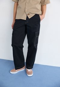 Black cargo pants with side pockets, tan stitching, and a relaxed fit, paired with light brown lace-up shoes on a blue surface.