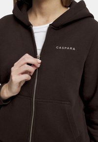 Svart zip-up hettegensere med frontglidelås, sidelommer, og hvit logo "CASPARA" trykt på brystet. Glatt stofftekstur.