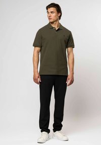 Muž stojící, oblečený v olivově zelen�ém polo tričku, černých kalhotách a bílých teniskách, dívající se na stranu na pozadí jednoduchého světlého pozadí.