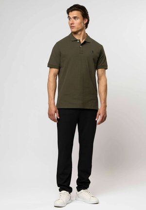 Mies seisoo, päällä oliivinvihreä polo-paita, mustat housut ja valkoiset tennarit, katsoo sivulle yksiväristä vaaleaa taustaa vasten.