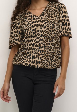 Frau trägt eine kurzärmelige V-Ausschnitt-Bluse mit Leopardenmuster und schwarze Hose, steht vor einem einfachen hellen Hintergrund.