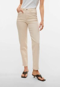 Lysebeige jeans med slim fit, fem lommer, subtil søm og høy midje, paret med svarte flip-flops.