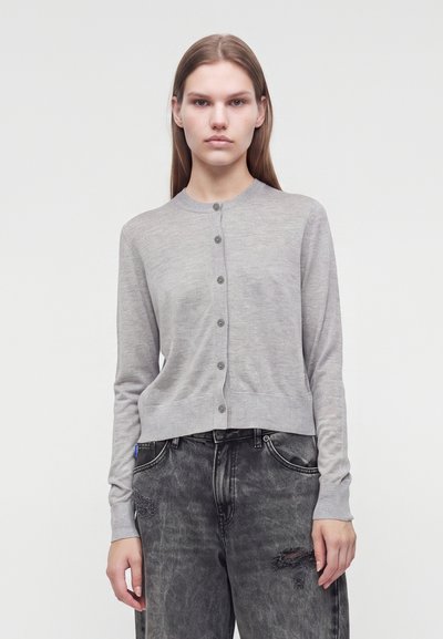 Jeune femme face à l'appareil portant un cardigan gris clair boutonné et un jean noir déchiré sur fond blanc uni.