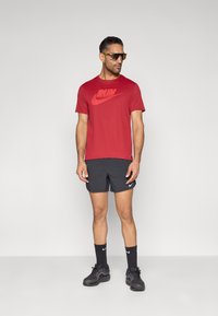 Κόκκινο μπλουζάκι Nike με το λογότυπο «RUN», σε συνδυασμό με μαύρο σορτς. Φοράει αθλητικά μαύρα αθλητικά παπούτσια και γυαλιά ηλίου, στέκεται μπροστά σε μια απλή φόντο.