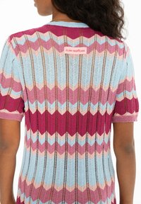 Dzianinowy top z krótkimi rękawami w zygzakowaty wzór, w odcieniach różowego, niebieskiego i bordowego; z małą metką z napisem "CLUB TROPICANA" z tyłu.