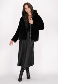 Sort faux fur jakke med hætte, lynlås foran og lange ærmer, parret med en sort satin nederdel og knæhøje sorte stiletto støvler.
