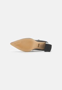 Chaussure à talon haut noire avec un bout pointu, une tige en matériau lisse, une semelle beige texturée et un talon carré. Logo "GUESS" embossé sur la semelle.