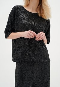 Top nero con paillettes, dalla vestibilità rilassata, maniche corte e scollatura rotonda. La texture presenta paillettes ravvicinate per un effetto luccicante.