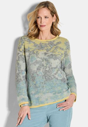 Femme aux cheveux blonds ondulés portant un pull à motifs pastel jaunes, bleus et gris et un pantalon bleu clair, se tenant avec une main sur la hanche.