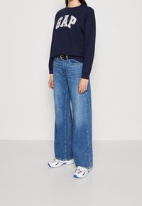 Sudadera azul marino con el logo "GAP", combinada con vaqueros de pierna ancha en un azul claro y zapatillas blancas con detalles azules y grises.