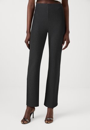 Pantalon noir à taille haute à fines rayures porté avec des sandales noires à talons hauts ouvertes sur une personne debout sur un fond uni.