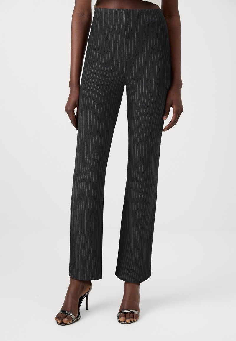 Pantalon noir à taille haute à fines rayures porté avec des sandales noires à talons hauts ouvertes sur une personne debout sur un fond uni.