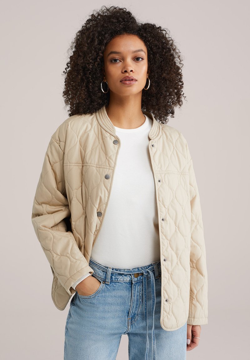 Veste bomber matelassée beige avec boutons-pression, col côtelé et coupe décontractée. Associée à un haut blanc et un jean bleu clair.