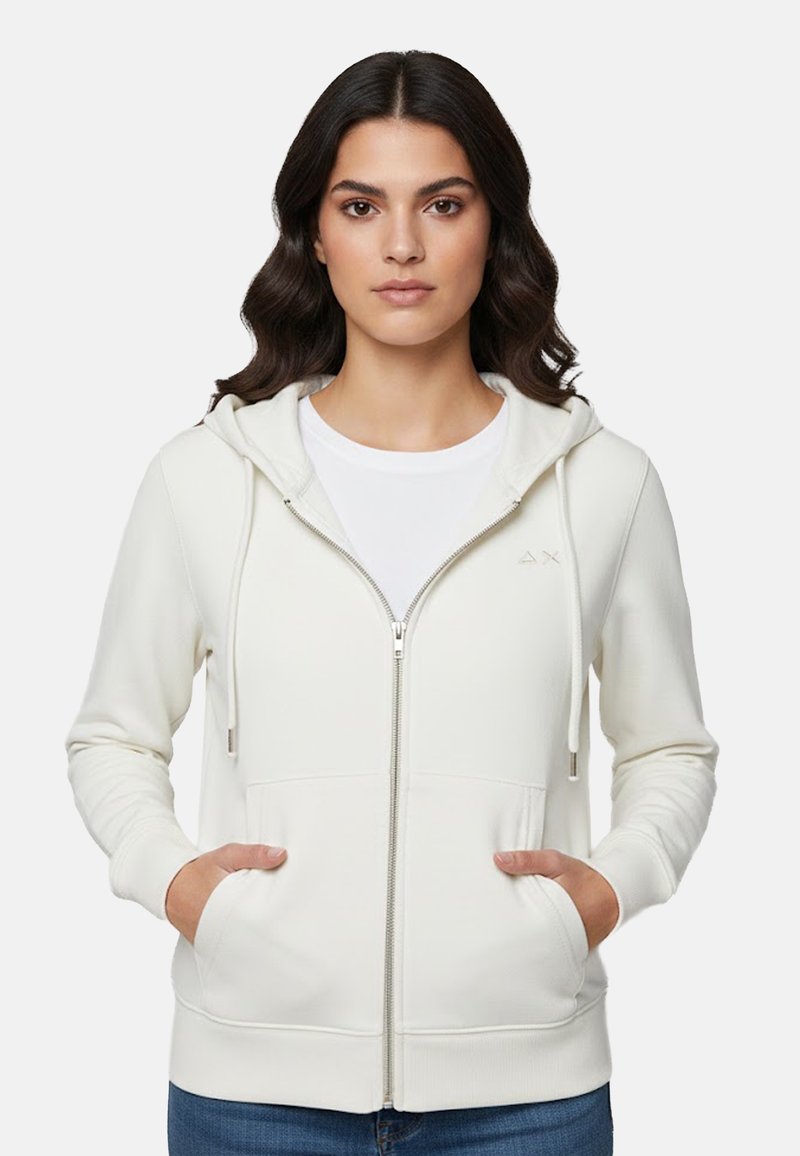 Giovane donna con felpa bianca con zip e mani nelle tasche anteriori, abbinata a jeans blu, sfondo neutro.