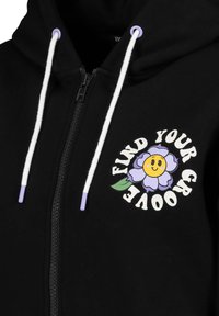Zwarte hoodie met rits, voorzien van witte trekkoorden en paarse accenten, versierd met een bloemenafbeelding en de tekst "VIND JOUW GROOVE."