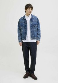 Jeans jakna z dvema prsne žepi, bela srajca z gumbi spodaj, temne hlače in temni usnjeni čevlji. Model stoji na svetli podlagi.