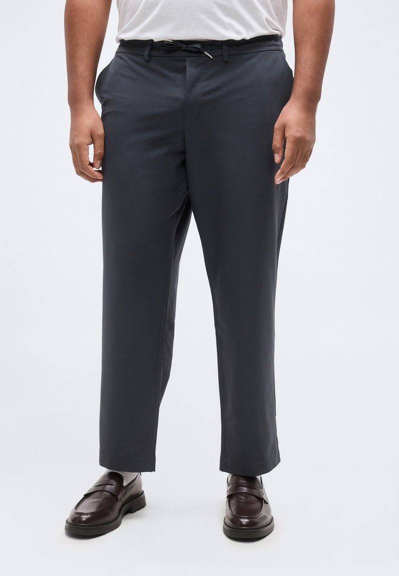 Άνδρας που φοράει σκούρα γκρι παντελόνια προσαρμοσμένης ραφής με κορδόνι στη μέση και σκούρα καφέ δερμάτινα μοκασίνια μπροστά σε λευκό φόντο.