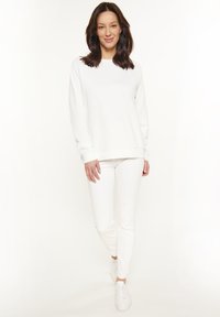 Felpa bianca con un taglio rilassato, polsini e orlo a costine. Abbinata a pantaloni bianchi slim-fit e scarpe da ginnastica bianche, su uno sfondo chiaro.