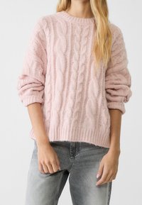 Jasnoróżowy sweter w splot kablowy z okrągłym dekoltem, oversize'owymi rękawami i ściągaczem na dole. Noszony z jasnoszarymi dżinsami.