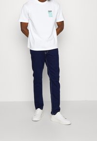 Bílé bavlněné tričko s malou tyrkysovou grafikou na levé hrudi, doplněné tmavě modrými džínami a bílými teniskami.