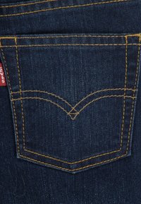 Tumši zila džinsa bikses ar aizmugurējo kabatu, kurai ir dzeltējoša šuve. Kabatas raksturo liekta forma ar izteiktu V veida simbolu un sarkanu zīmola etiķeti.