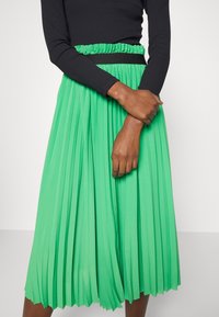 Jupe plissée verte avec une taille élastique et des accents noirs. Fabriquée en tissu léger, elle présente des lignes de pli verticales pour une texture.