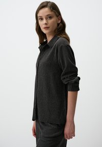 Zwarte gestructureerde button-up shirt met een relaxte pasvorm, voorzien van een kraag en opgerolde mouwen, weergegeven tegen een witte achtergrond.
