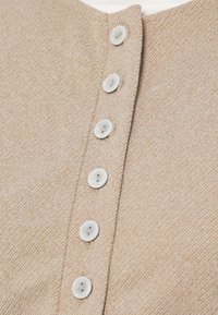 Beige kofta med ett strukturerat tyg, utrustad med sex runda vita knappar längs framkanten. Mjukt, diskret stickmönster synligt.