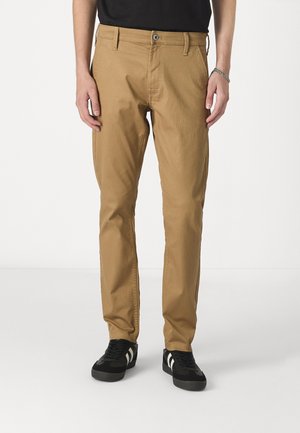 Tan chinos featuring a classic fit, belt loops, and a single button closure. Paired with black shoes with white accents.