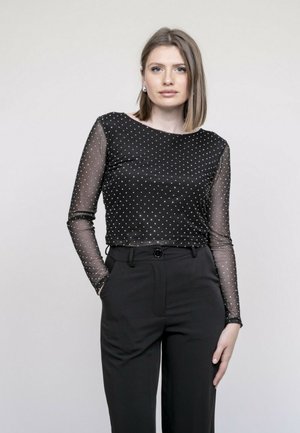 Jeune femme aux cheveux blonds raides portant un haut noir transparent à manches longues avec des pois blancs et un pantalon noir taille haute, debout.