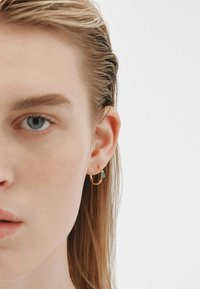 Pendiente de aro dorado con un pequeño acento en turquesa, presentado sobre un fondo claro y neutro. El cabello está lacio y mojado, dejando ver el pendiente.