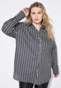 Camisa de botones a rayas blancas y negras, con mangas largas, cuello y corte relajado. Tejido suave, con botones blancos y dobladillo más largo.