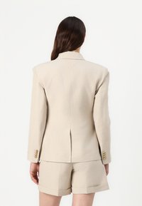Beige skreddersydd blazer med en enkelt ventil bak og tre gullknapper på hver mansjett, kombinert med matchende shorts.