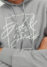 Sudadera de algodón gris con un gran estampado gráfico blanco y texto en cursiva. Incluye capucha ajustable con cordones a juego y puños acanalados.