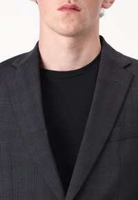 Tamsiai pilkas bleizeris su subtiliu langelių raštu, notch tipo atlapais ir vienu krūtiniu kišeniu, dėvimas ant juodos marškinėlių.